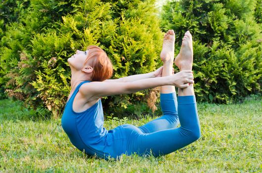 Beautiful red woman doing fitness or yoga exercises outdoors in a green park