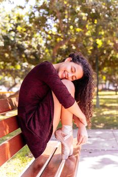 Young beautiful ballerina in black dress sitting on a bench in a park with her eyes closed and smiling