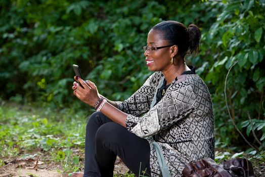 Beautiful woman resting in the forest speaks by mobile phone sitting at the park in casual clothes. Close up of big greenery in the park and holding the smart phone. technology and communication concept