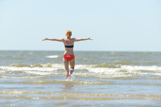 Slim young woman running into sea waves with outstretched hands