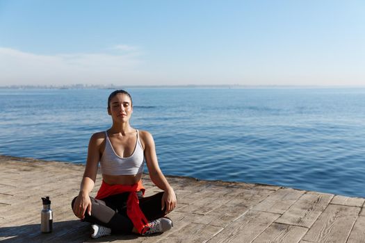 Outdoor shot of peaceful fitness woman sitting in asana and doing yoga near the sea. Female athlete meditating on seaside.