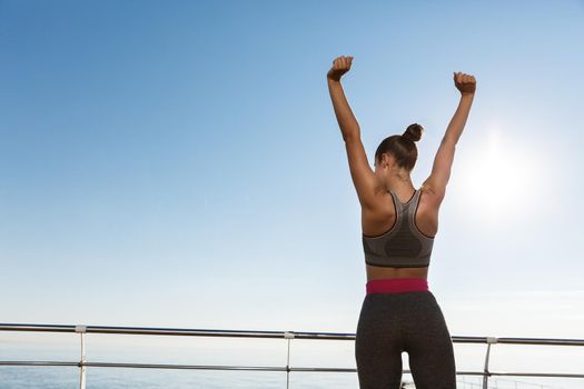 Rear view of happy triumphing sportswoman standing on a pier, raising hands up like champion, achieve goal during training session outdoors.