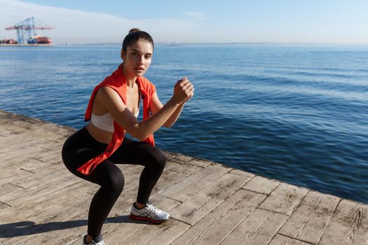 Outdoor shot of attractive fitness woman workout near the sea, doing squats exercises and looking at camera.