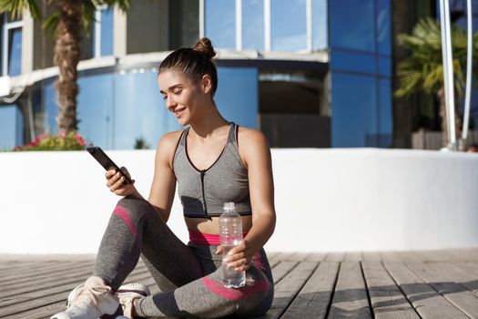 Outdoor shot of happy attractive sportswoman sitting on floor after workout, drinking water and looking at smartphone.