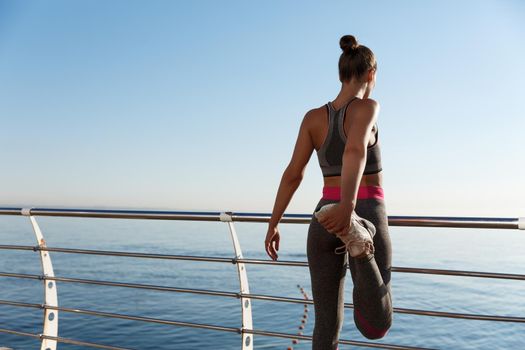 Rear view of attractive fitness woman stretching her legs before jogging, leaning on handrail along seaside promenade, training outdoors.