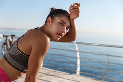 Close-up of attractive sportswoman taking a breath during workout, looking at camera and wiping sweat off forehead, jogging along seaside promenade.