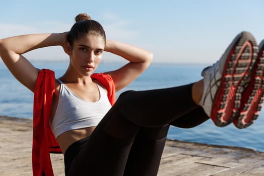 Close-up of confident young fitness woman working out on seaside promenade, doing crunches exercise on pier at daytime, looking at camera.