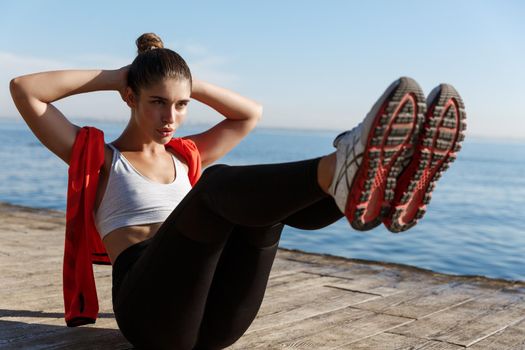 Young confident sportswoman doing crunches on a pier, training abdominal abs, working out on seaside promenade.