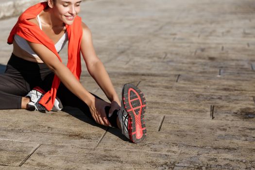 Cropped shot of attractive fitness woman stretching leg and smiling, doing warm-up exercises before running.