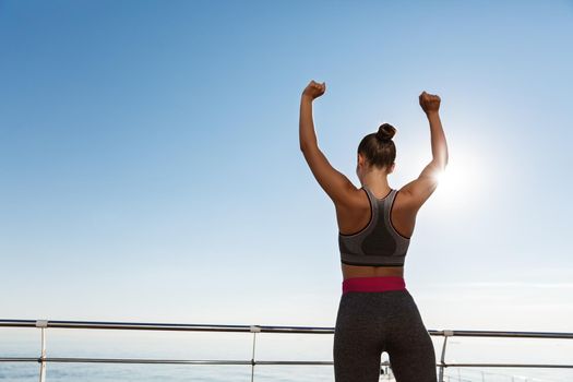 Rear view of happy and satisfied fitness woman looking at the sea and raising hands up like champion, achieve goal during workout or jogging on the seaside promenade.