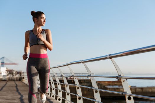 Portrait of young healthy woman jogging on the seaside promenade, running and looking at the sea thoughtful. Runner workout outdoors, training alone.