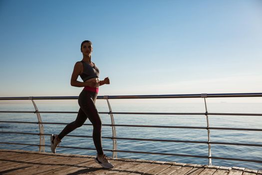 Side view of young fitness woman running along the pier. Sportswoman training on seaside promenade.