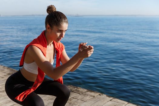 Side view of focused young sportswoman doing squats near the sea, having training on seaside.