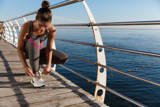 Outdoor shot of attractive sportswoman tying shoelaces on a pier, jogging at seaside.