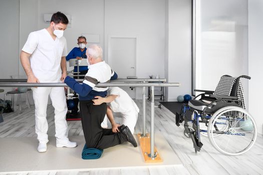 Physiotherapists helping disabled Caucasian senior man to walk with parallel bars in rehabilitation center. High quality photo