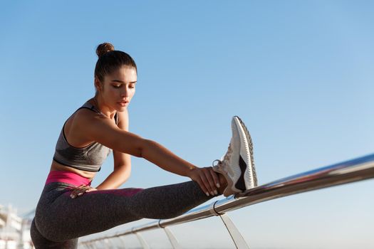 Image of attractive fit fitness girl workout on a pier. Sportswoman stretching legs and warming-up before jogging training on the seaside promenade.