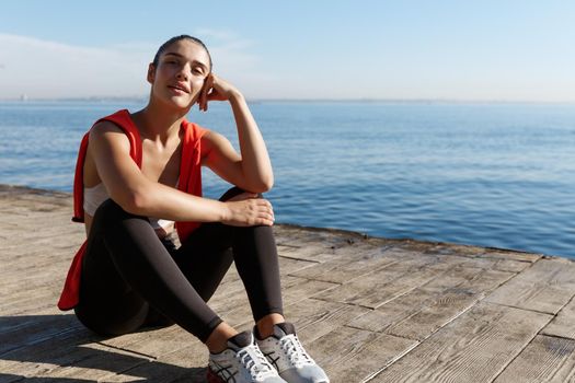 Outdoor shot of satisfied fitness woman sitting on seaside promenade and looking at the sea, finish workout and having a break.