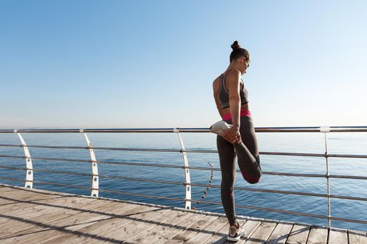 Rear view of attractive fitness woman stretching her legs before jogging, leaning on handrail along seaside promenade, training outdoors.