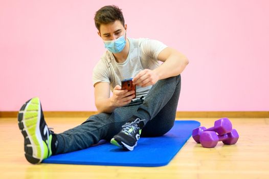 Fitness man Relaxing after training using mobile phone, wearing protective face mask during Covid 19 new normal. High quality photo