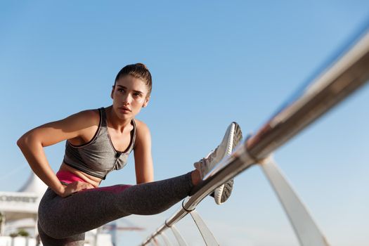 Low angle shot of attractive fit woman workout on a pier, stretching her leg before jogging training. Girl warming-up for morning run, holding hand on a handrail.