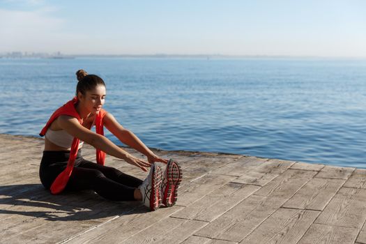 Outdoor shot of beautiful fitness woman stretching and training near sea, reaching toes with hands to warm-up before workout.