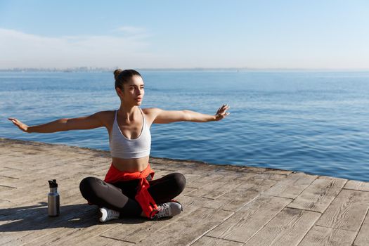 Outdoor shot of relaxed young woman sitting on wooden pier and looking at the sea, doing yoga exercises.