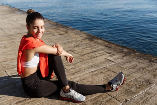 Outdoor shot of smiling fitness woman sitting near the sea and looking happy, workout on seaside promenade.