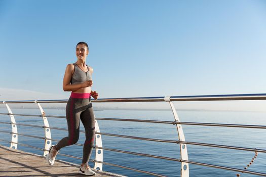 Image of happy fitness woman jogging along seaside promenade, smiling during workout.