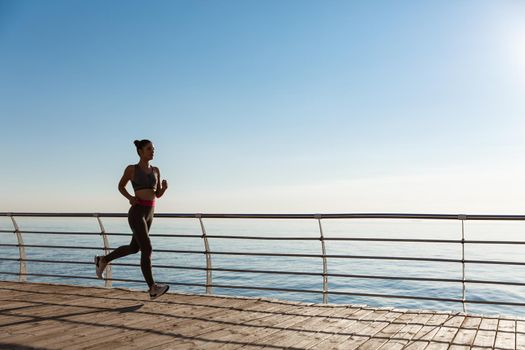 Side view of young fitness woman running along pier. Female athlete workout and jogging on the seaside promenade.