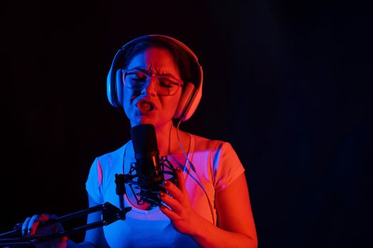 Caucasian woman in glasses and headphones sings into a microphone in neon light on a black background. An emotional girl is recording a song in a recording studio.