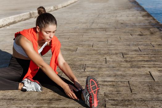 Outdoor shot of young fitness woman training on seaside promenade, sitting on pier and stretching leg before workout.