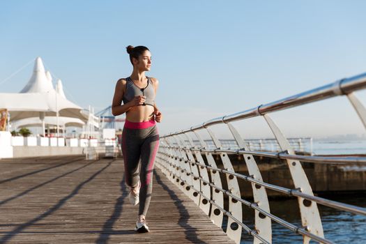 Full length of attractive fit woman jogging in the morning, looking at sea. Female runner working out on the seaside promenade, running towards camera.