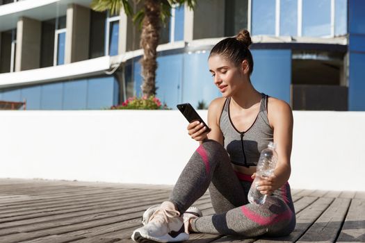 Outdoor shot of attractive fitness woman sitting on wooden pier with smartphone, having break after workout, drinking water.