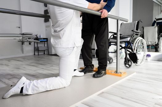 Physiotherapist helping disabled Caucasian senior man to walk with parallel bars in rehabilitation center. High quality photo