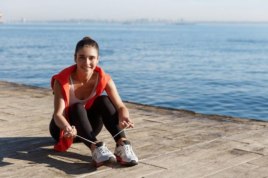 Outdoor shot of attractive smiling sportswoman sitting on pier and tying shoelaces during workout, training near the sea.