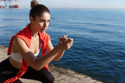 Close-up of focused young female athlete workout on a pier, doing squats and looking at sea.