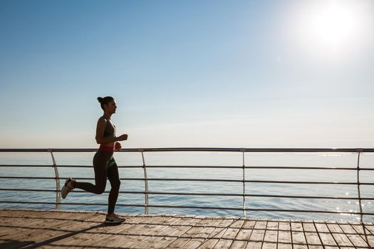 Side view of young fitness woman running along pier. Female athlete workout and jogging on the seaside promenade.