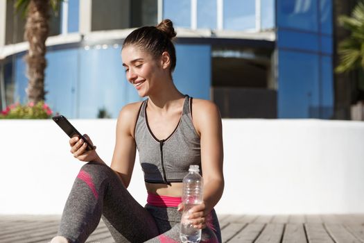 Outdoor shot of happy fitness woman looking at mobile phone app, sitting on wooden floor and drinking water.