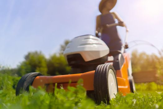 A young girl is mowing a lawn in the backyard with an orange lawn mower. A woman gardener is trimming grass with the grass cutter, bottom view.A lawnmower is cutting a lawn on a summer sunny day.