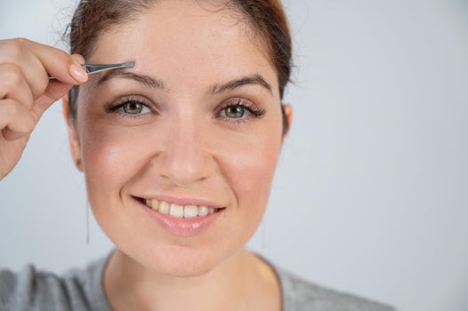 Close-up portrait of a caucasian woman doing eyebrow correction herself with tweezers.