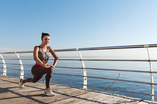 Outdoor shot of smiling fitness woman stretching and working out on the seaside promenade.