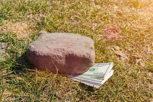 A large bundle of money lies on the ground, pressed against the grass by a stone