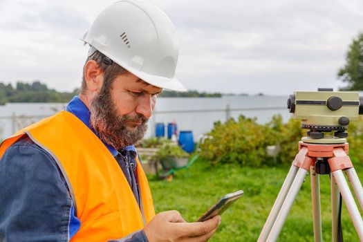 A civil engineer with an optical level looks into a mobile phone. A bearded man is looking for information on his mobile phone.