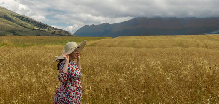 Happy young woman in floral dress holding her hat with her hands walking alone in the middle of a wheat field on a cloudy day with blue sky and mountains in the background