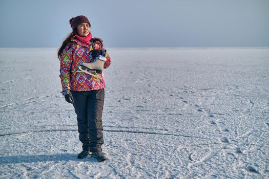 A woman in a ski suit holds skates on her shoulder and looks into the distance, after skiing on a frozen lake