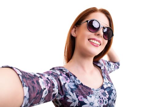 Beautiful smiling young girl with sunglasses taking a selfie, looking at the camera POV, isolated on white background. Attractive redhead girl with long hair taking a selfie. Lifestyle, happiness, and social concept.