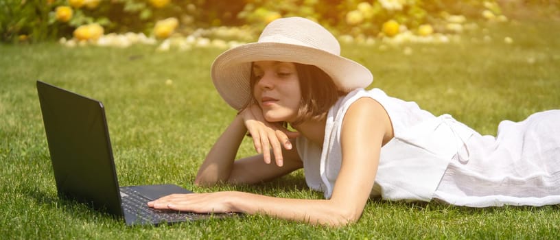 A young girl in a white dress is working on a laptop, texting on the Internet on her device, freelancing, studying, resting on a meadow in a park, lying on the grass.