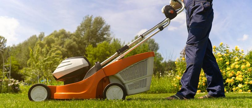 A young man in a straw hat is mowing a lawn with a lawn mower in his beautiful green floral summer garden. A professional gardener with a lawnmower cares for the grass in the backyard.