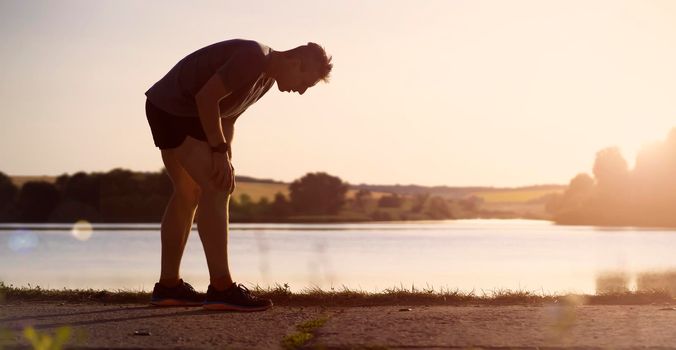 A young man jogging at sunset by the lake, the tired athlete holds on to his sore knee.