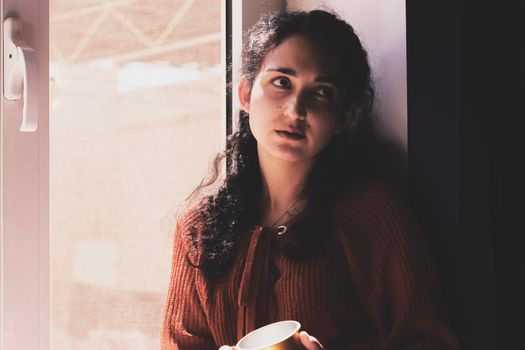 Cold autumn days - a young multi-racial female drinks coffee in a cozy windowsill. Middle-eastern or mixed-race woman enjoys weekend drinking hot tea near the windows